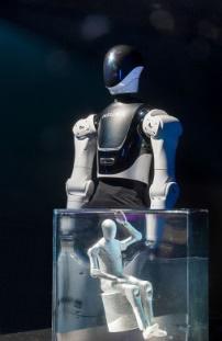 Ein Roboter sitzt in einer Glasbox, während ein Mann daneben auf dem Boden sitzt, wodurch eine einzigartige Interaktion zwischen ihnen entsteht.