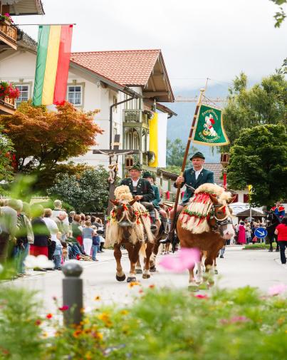 Eine Gruppe von Menschen reitet auf Pferden eine Stadtstraße entlang, umgeben von Gebäuden und Zuschauern.