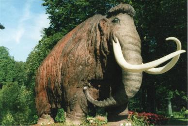 A large woolly mammoth statue stands prominently in the center of a park, surrounded by green grass and trees.